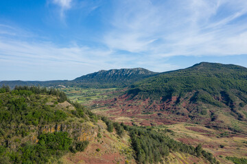 Expansive aerial landscape featuring striking red earth hills and lush green forest under a wide blue sky with wispy clouds. The terrain feels rugged, open, and naturally dramatic.