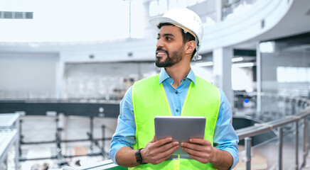 A man in a hard hat and safety vest stands in a construction zone. He looks around while holding a tablet and seems to monitor the work being done. The setting is bright and spacious.