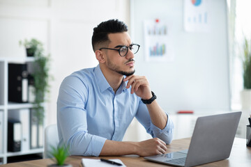 Troubled Arab businessman sitting at his desk, looking at laptop screen, solving work problem in...