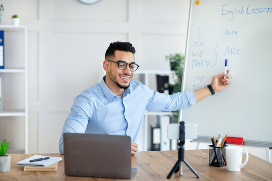 Cyberlearning, remote education concept. Happy Arab male teacher giving online English lesson, using smartphone and laptop, pointing at blackboard with grammar rules at home office