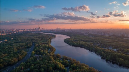 Fototapeta premium Aerial view of a city beside a river, with trees and buildings. The sky is painted with sunset colors and scattered clouds