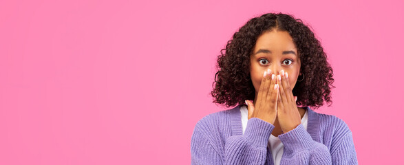 A young person stands in front of a pink background with hands covering their mouth. They have...