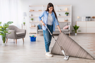 Fototapeta premium Household Concept. Full length of smiling lady washing hardwood laminate flooring using water spray mop pad, cleaning under the carpet in living room. Blurred Background, Free Space, Modern Interior