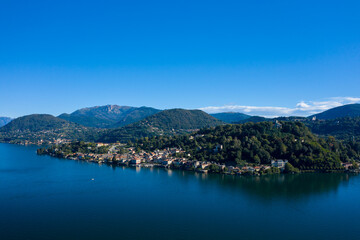 Obraz premium Sweeping view of a lakeside town nestled at the foot of densely forested hills along Lake Orta in northern Italy. The tranquil blue water reflects the surrounding greenery and distant mountain slopes