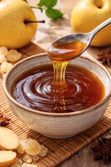 Golden honey drips from a spoon into a bowl, encircled by ripe pears, glistening sugar crystals, and fragrant star anise on woven mat.