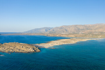 Aerial perspective of a rocky headland and calm blue lagoon at Elafonisi beach in Crete, Greece, with distant rugged mountains and a sunlit, cloudless sky. The landscape feels open, textured, and