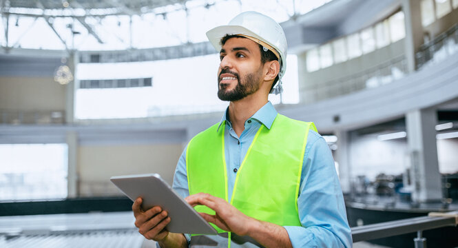 A construction worker stands inside a building site wearing a hard hat and safety vest. He looks at a tablet as he manages tasks and checks information.