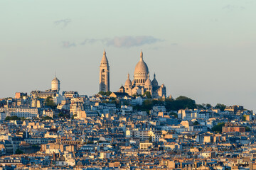 Naklejka premium Distant view of the Sacre Coeur basilica crowning Montmartre hill, surrounded by layered Parisian rooftops glowing in soft golden evening light under a clear sky.