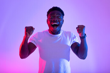 A young African American male expresses pure joy and triumph, shouting in excitement and gesturing with his fists raised in a vibrant neon-lit space, celebrating a significant achievement.