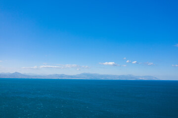 Vast expanse of deep blue water stretching toward a hazy mountain range on the horizon, beneath a clear sky with scattered clouds. The scene feels open, calm, and filled with natural light.