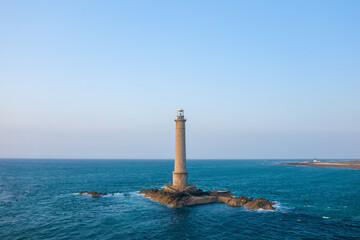 A tall stone lighthouse rises from a rocky islet surrounded by vivid blue sea at Cap de la Hague, under a clear sky. The tranquil scene features open water and distant coastline, evoking maritime