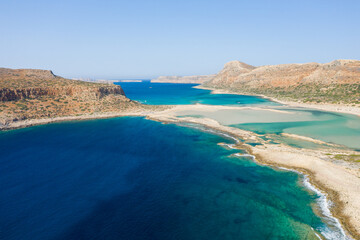 A sweeping aerial perspective reveals the vivid blue and green waters of Balos Beach lagoon in Crete, Greece, bordered by rocky hills and sunlit sandy stretches. The landscape feels open, natural, and