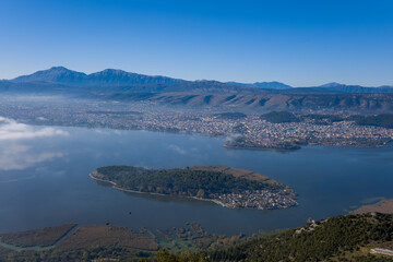 Elevated perspective reveals the tree-covered island of Ioannina set in tranquil lake waters, with the city sprawling along the shore and the Pindus mountains rising in the background. Crisp blue sky