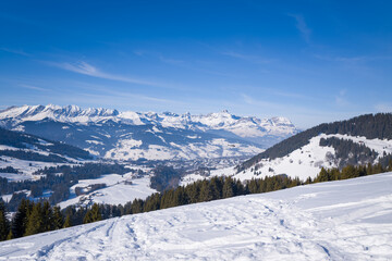 Wide winter view over snow-blanketed hills, pine forests, and the Megeve valley, with dramatic mountain peaks rising under a vivid blue sky. Crisp sunlight highlights the untouched snow and creates a