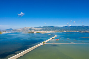 Long causeway stretches across the vivid blue and green waters of the Missolonghi salt lagoon in western Greece. The scene features distant mountains, a clear sky, and geometric lines of salt marshes