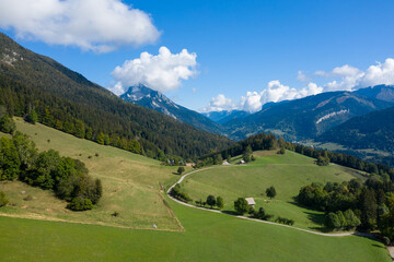 A curving country road weaves across lush green meadows and forested slopes in the Chartreuse Massif, with dramatic peaks and scattered clouds under a vibrant blue sky. The scene highlights the
