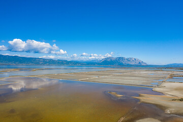 Geometric salt pans stretch across the Missolonghi wetland in western Greece, with clear reflections and a distant mountain range under a vivid blue sky. The expansive landscape features tranquil
