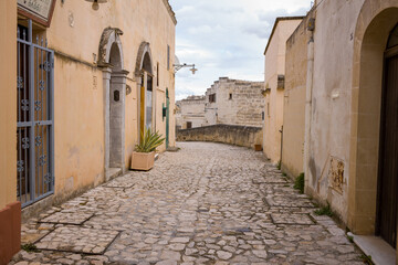 A peaceful, empty cobblestone street winds between weathered stone buildings in Matera, Italy. Soft daylight highlights textured walls and rustic architectural details, creating a serene Mediterranean