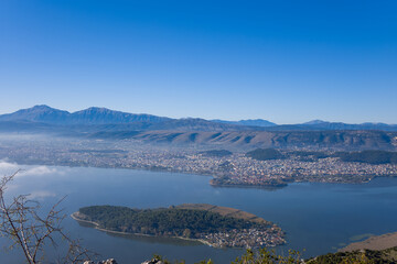 Elevated panoramic view of the Island of Ioannina surrounded by calm lake waters, with the city of Ioannina and the rugged Pindus mountains forming a dramatic backdrop in Epirus, Greece. Clear blue