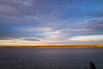 Soft golden light highlights the distant marsh grasses along the horizon of a tranquil Camargue lagoon, beneath a dramatic sky with layered blue and lavender clouds.