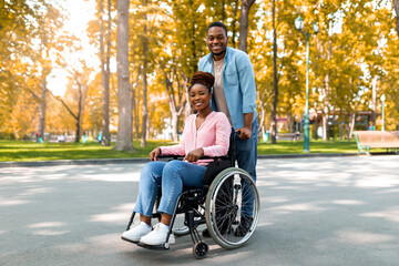 Full length of cheerful black guy taking his impaired girlfriend in wheelchair for walk at city...