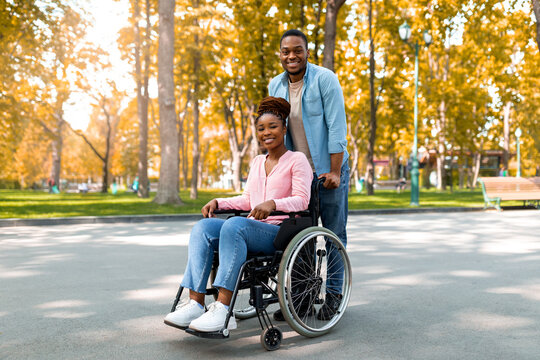 Full length of cheerful black guy taking his impaired girlfriend in wheelchair for walk at city park in autumn. African American disabled couple enjoying fall nature together, outdoors