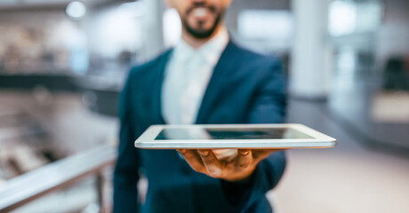 A businessman holds a tablet out in front of him in a modern office setting. The background...