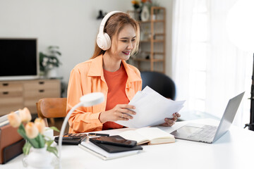 Smiling Asian woman wearing headphones and working on laptop at home office, happy student or freelancer enjoying online learning.