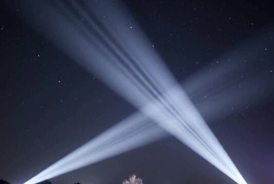 Two powerful searchlights illuminate starry night sky with dynamic light beams creating captivating X-shaped patterns and elegant sweeping movements, cinematic footage