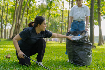 Two Asian women are picking up litter on the lawn in a park, participating in a daytime environmental cleanup.