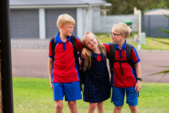 Three happy primary aged kids with backpacks standing together outside home before school