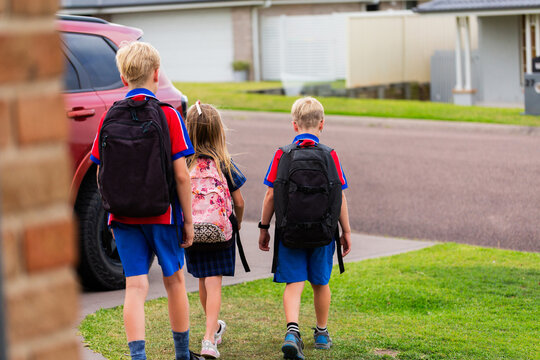 Three kids walking away from home to school bus stop carrying backpacks