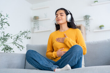 Happy young Asian woman practicing yoga and meditation at home sitting on couch in living room in lotus position and relaxing with closed eyes. Mindful meditation and wellbeing concept