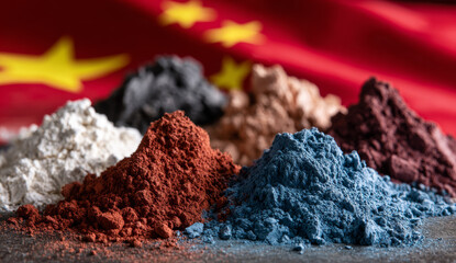 Colorful piles of finely textured powder pigments in various shades including white, black, red, blue, and brown, arranged on a dark surface with a blurred red and yellow flag in