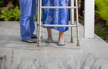 Senior woman practicing walking with aluminum walker and caregiver support in nursing home garden