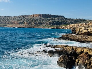 Dramatic cliffs overlooking the turquoise, turbulent sea at Cape Greco Peninsula, Cyprus, with waves crashing against rocky shores under a partly cloudy sky