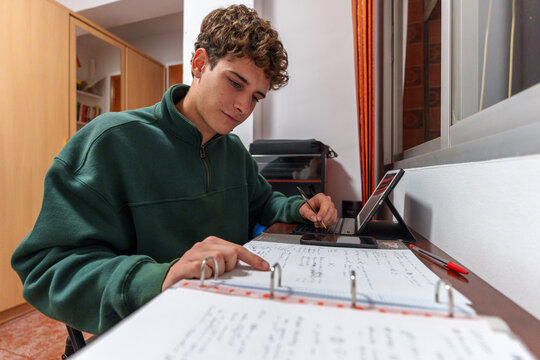 Young student learning and studying at desk, concentrating on homework with notes and technology indoors