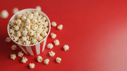 A red-and-white striped popcorn bucket overflowing with fluffy popcorn kernels on a red surface