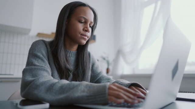 Freelance journalist or web-developer working in home workplace, female portrait . African american woman sitting at table with modern laptop and thinking, typing message and sending email to clients