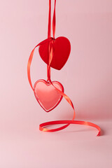 A pair of red hearts hang on red bow on a soft pink background, representing eternal love, warmth, and romance.