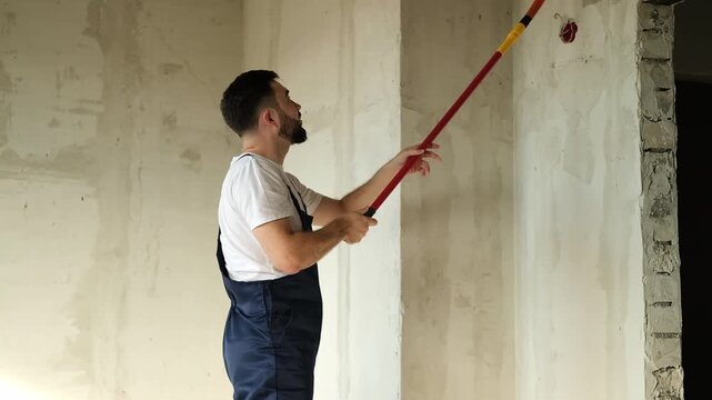 A bearded man in a work uniform is priming the walls after applying gypsum plaster. 