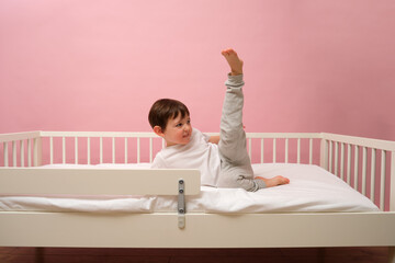 Happy little boy in white t-shirt and grey pants lying on white mattress in bed with one leg raised up. Child playing in bedroom. Child aged 4 years (four years old)