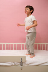 Happy kid boy jumping on white bed. Energetic child playing in bedroom. Childhood joy and playful activity at home concept. Child aged 4 years (four years old)