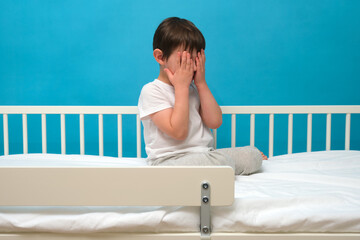 Child hiding face on bed. Boy playing hide and seek. Kid sitting with hands covering eyes on simple white bed against blue wall for early childhood and play. Child aged 4 years