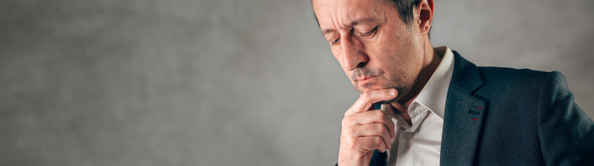 Thoughtful businessman looking down with hand on chin, expressing doubt, concern, decision making, uncertainty, or problem solving in a business context.