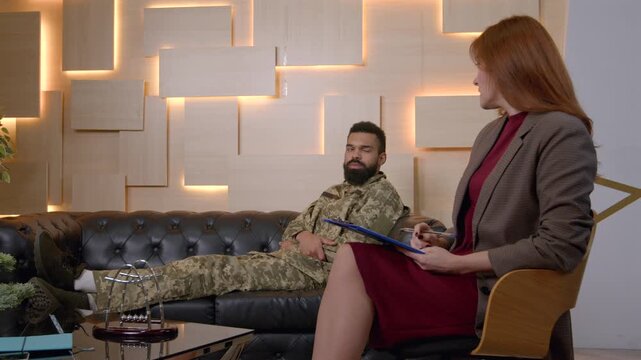 Middle aged woman psychotherapist and relaxed African American male army soldier in military, uniform on sofa, talking about troublesome behavior and mental problems during psychotherapy session.