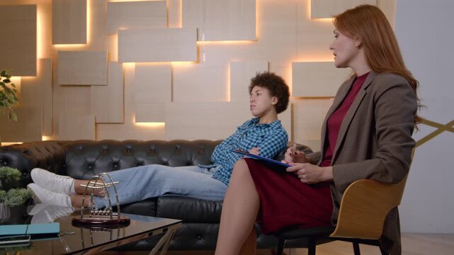 Charming mature female psychotherapist and stressed pretty African American woman on sofa, talking about troublesome behavior and mental problems during psychotherapy session.