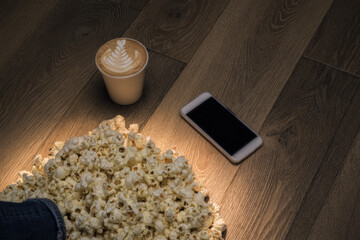 Cozy popcorn spill with latte and smartphone on wooden floor, warm light and relaxed mood