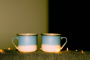 Two ceramic blue and white mugs with handles sit side by side on textured fabric lit by warm fairy lights, cozy minimal tabletop scene