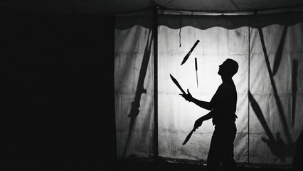 Man performing dangerous knife juggling circus act. Silhouette of juggler with knives on a white curtain. Entertainment show at chapiteau.